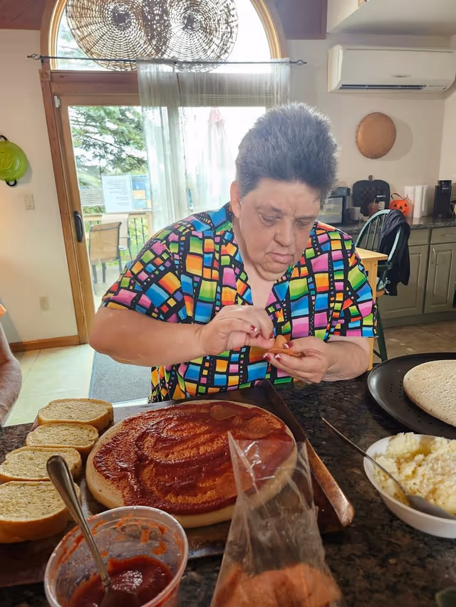A woman in a colorful patterned shirt is preparing a pizza in a kitchen. She is placing pepperoni slices on a pizza crust that has tomato sauce spread on it. There are slices of bread, a bowl of cheese, and another pizza crust on the counter. The kitchen has green cabinets, a window with sheer curtains, and a door leading outside.