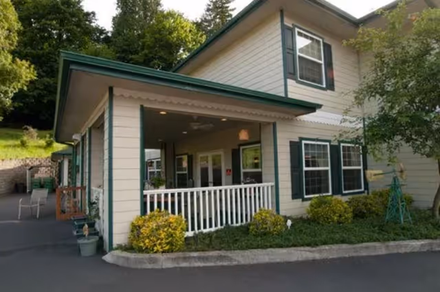 Exterior view of a two-story residential building with beige siding and green trim. The building has multiple windows with white frames and green shutters. There is a covered porch area with white railing and some outdoor seating. Shrubs and small plants are landscaped around the building, and trees are visible in the background.