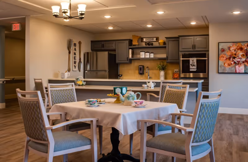 A dining area in a senior living facility featuring a square table with a white tablecloth, set with teacups, a teapot, and a cake stand. Surrounding the table are four cushioned chairs with wooden frames. In the background, there is a kitchen area with gray cabinets, a stainless steel refrigerator, an oven, and decorative signs labeled 'BAKERY' and 'GROCERY'. The room has warm lighting and a wooden floor.