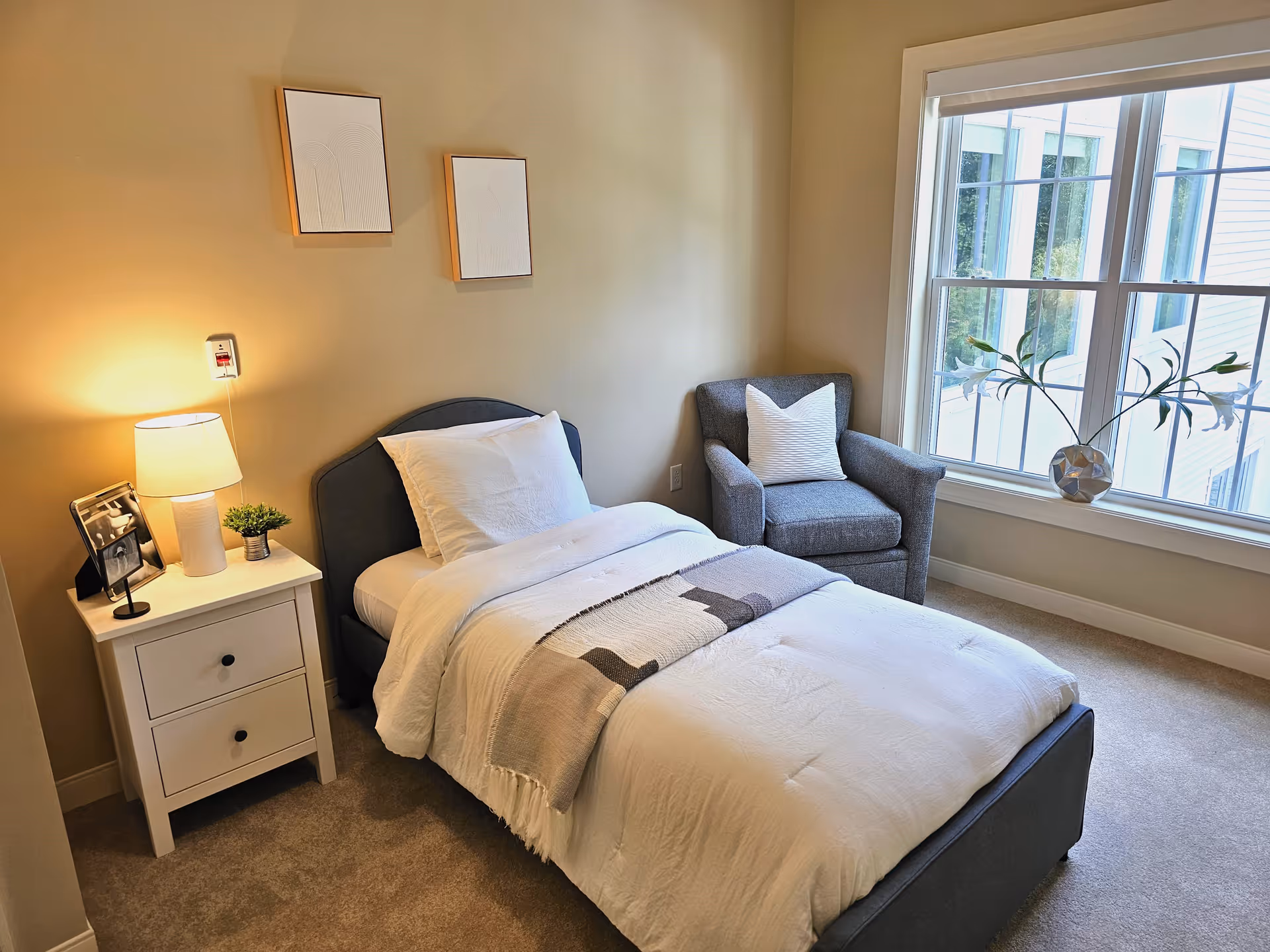 A cozy bedroom with a single bed made with white linens and a gray blanket. Next to the bed is a white nightstand with a lamp, a small plant, and a framed photo. A gray armchair with a white pillow is placed near a large window with a vase of flowers on the windowsill. Two minimalist framed artworks hang on the beige wall above the bed.