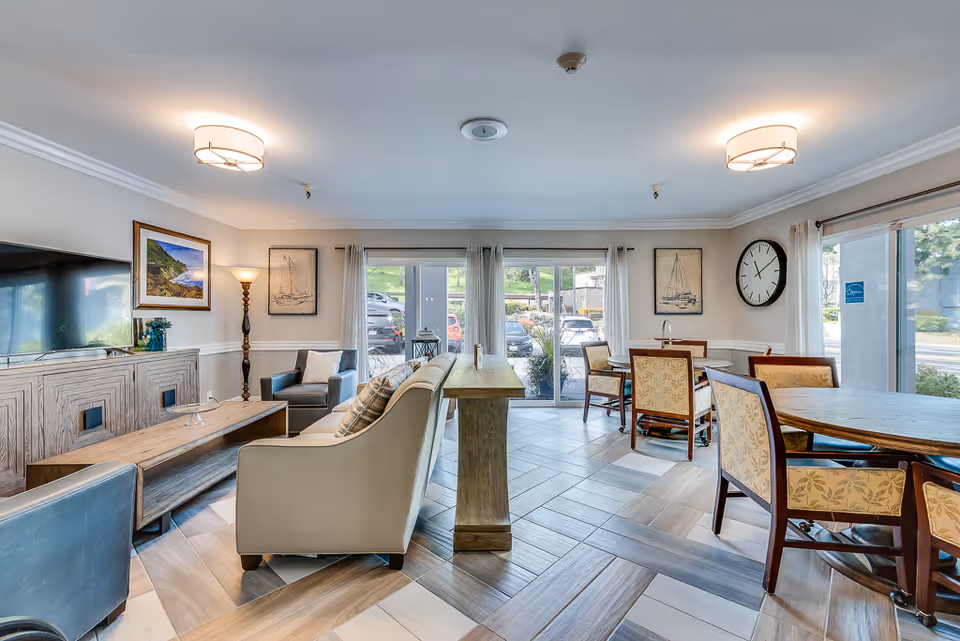 Bright communal lounge and dining area with sofas, armchairs, wooden tables, wall art, and large windows.