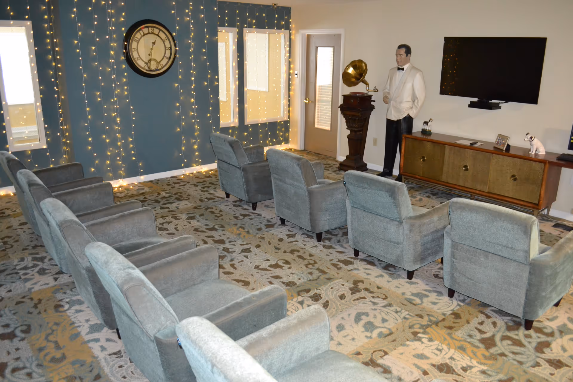 A cozy living room with two rows of gray upholstered armchairs facing a wall-mounted flat screen TV. The room features a patterned carpet, a large vintage-style clock on a blue accent wall adorned with string lights, and a life-sized statue of a man in a white tuxedo standing next to an antique gramophone on a wooden pedestal. A wooden console table beneath the TV holds decorative items including two dog figurines and a framed photo.