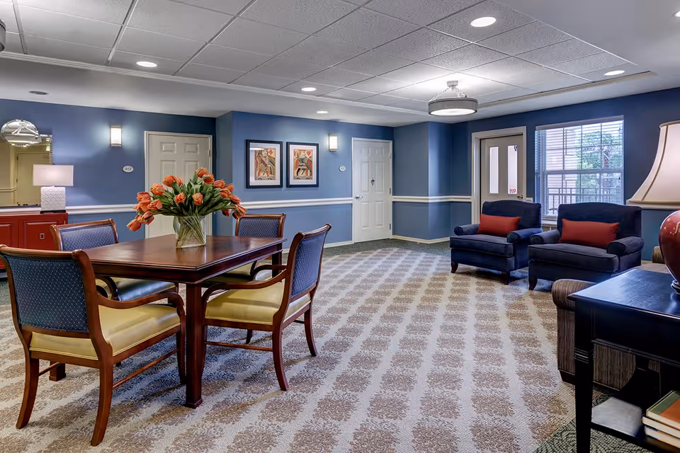A cozy senior living common area with a wooden table and four chairs, a vase with orange tulips on the table, two blue armchairs with red cushions near a window, blue walls with white trim, framed artwork, and a patterned carpet.
