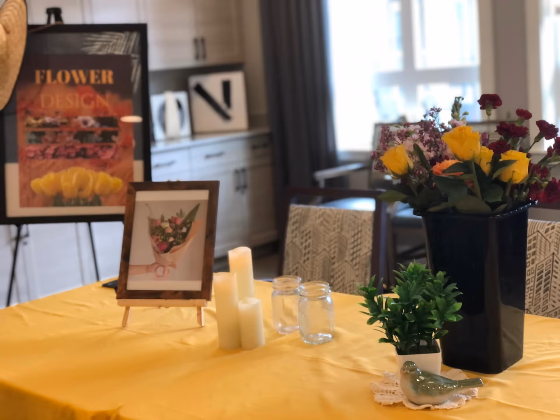 A table covered with a yellow tablecloth displaying a large black vase with a colorful bouquet of flowers, a small green potted plant, a ceramic bird figurine on a doily, three white pillar candles, and two empty glass jars. In the background, there is a framed picture of a flower bouquet on a small easel and a larger framed poster with the words 'FLOWER DESIGN' on an easel. The setting appears to be a bright room with white cabinets and a window with curtains.