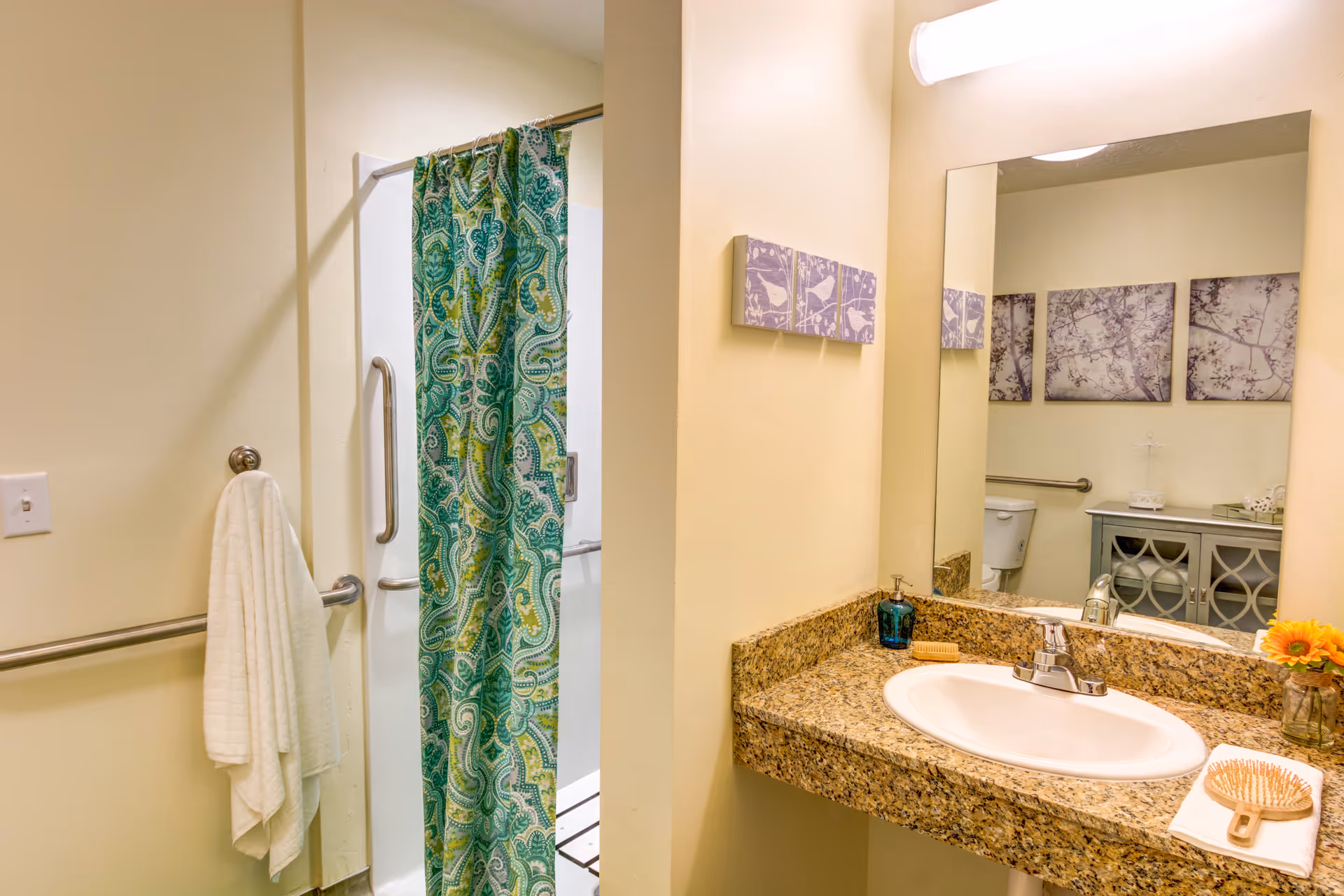 A bathroom with a granite countertop sink, a large mirror, and a shower with a green patterned curtain. A white towel hangs on a metal bar next to the shower. In the mirror's reflection, a toilet, a cabinet, and three framed pictures on the wall are visible. A small vase with yellow flowers and a hairbrush are on the countertop.