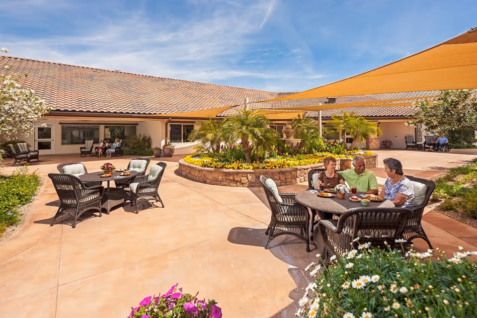 Outdoor patio area at Northbrook Inn Memory Care Community with people sitting at tables under a large shade sail. The patio features wicker chairs and tables, a central flower bed with palm trees and colorful flowers, and a building with a tiled roof in the background under a blue sky.
