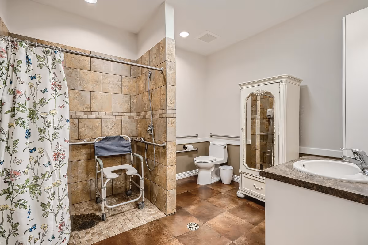 A spacious senior-friendly bathroom with a tiled walk-in shower featuring a floral shower curtain and a shower chair. The bathroom also includes a white toilet with grab bars, a white cabinet with a mirrored door, and a sink with a dark countertop. The floor is covered with large brown tiles.