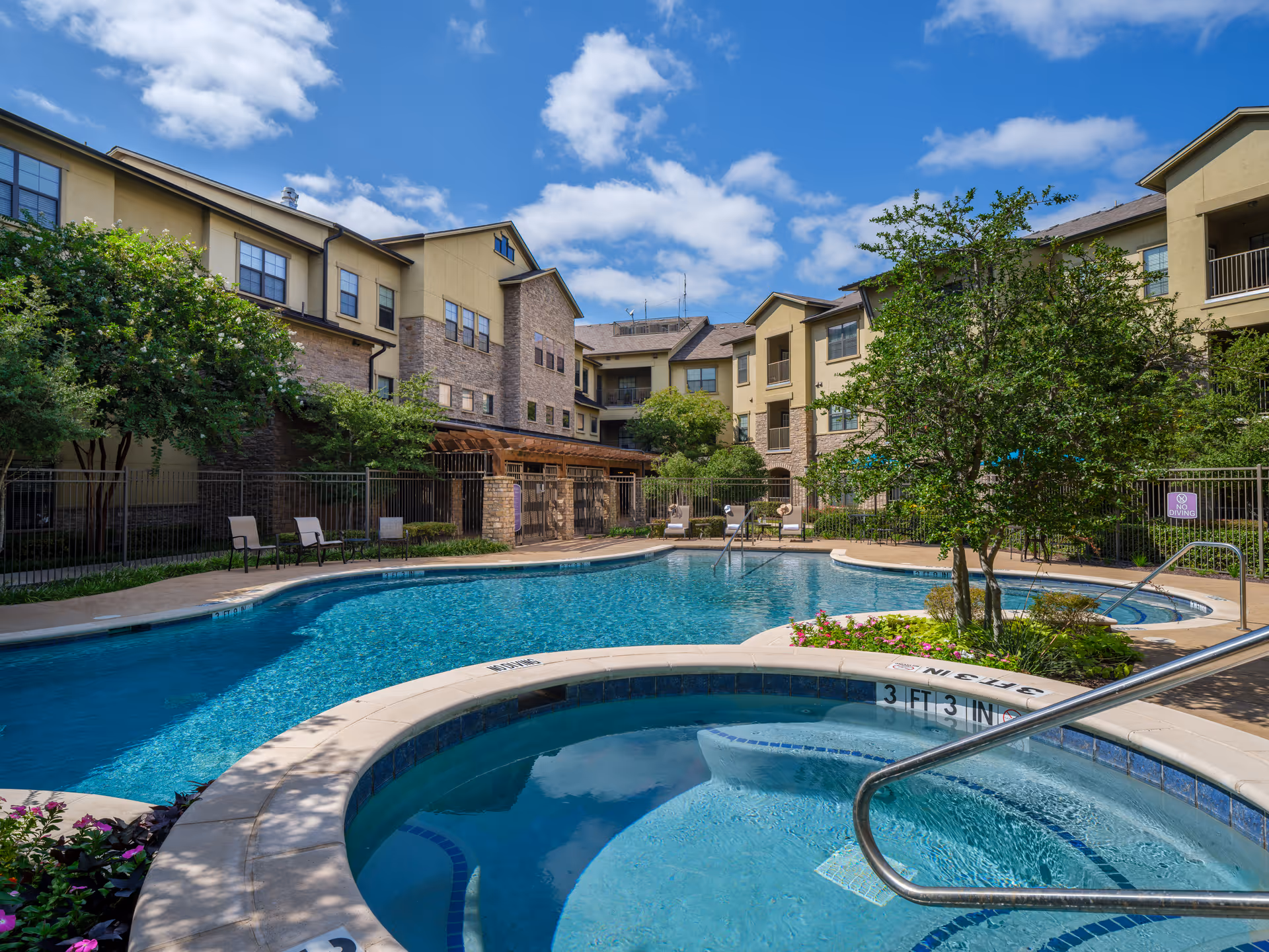 Outdoor swimming pool and hot tub area surrounded by a three-story residential building with balconies and windows. The pool area has lounge chairs, trees, and landscaping under a partly cloudy blue sky.