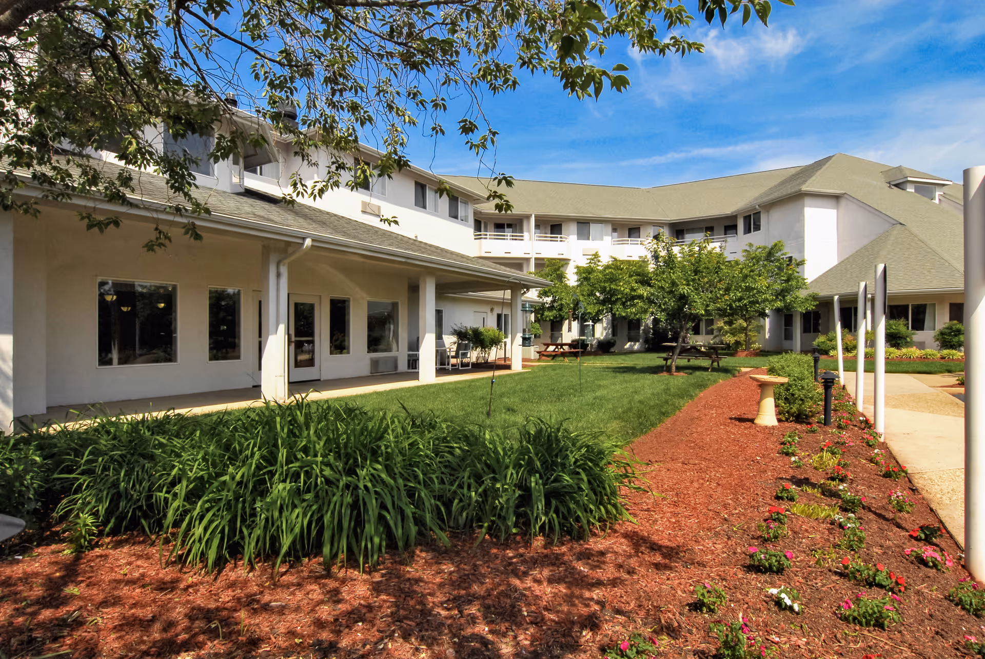A landscaped courtyard with a green lawn, flowerbeds, benches and a U-shaped white senior living building under a blue sky.