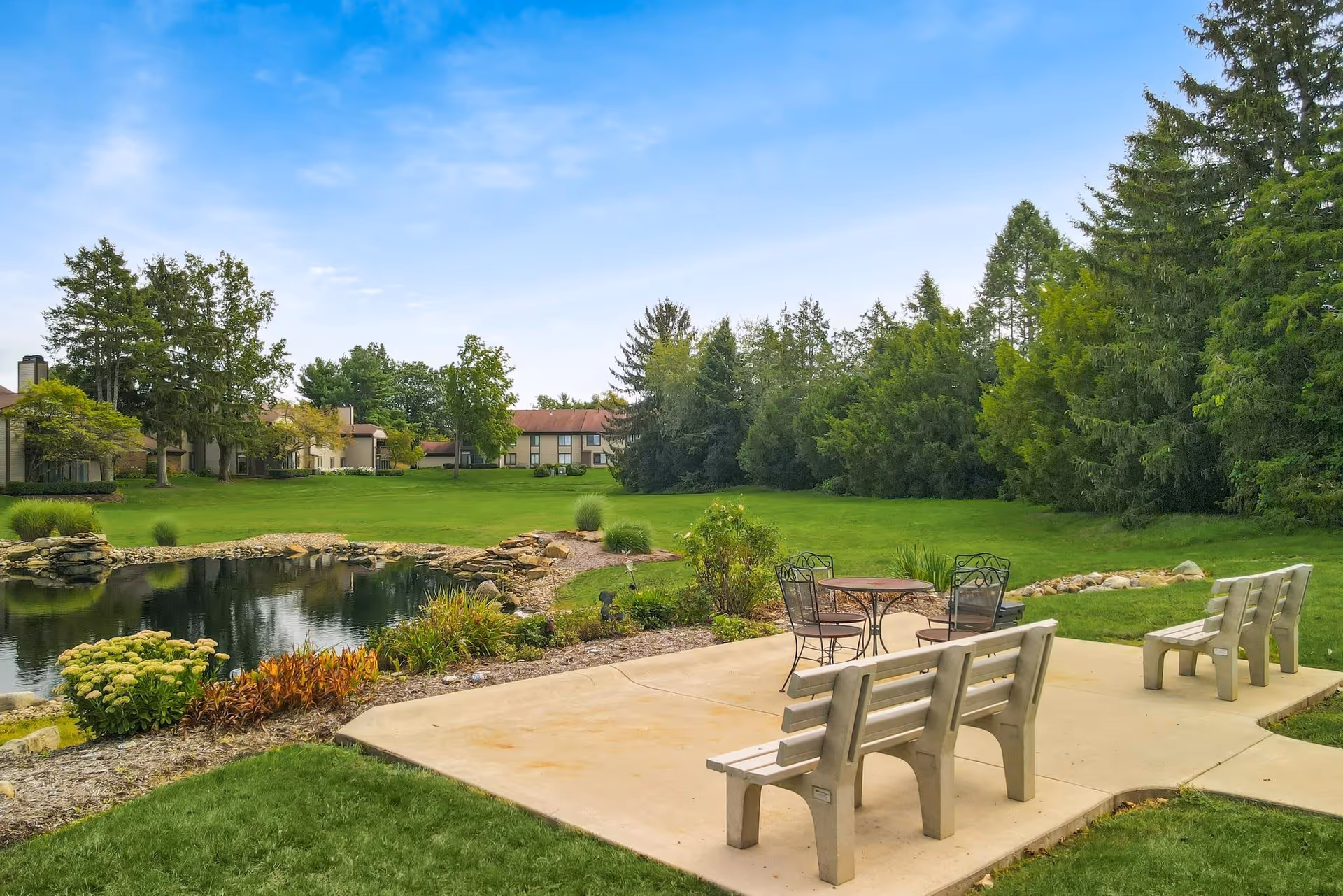Outdoor seating area with benches and a small round table with chairs on a concrete patio next to a pond surrounded by grass, trees, and shrubs under a blue sky.