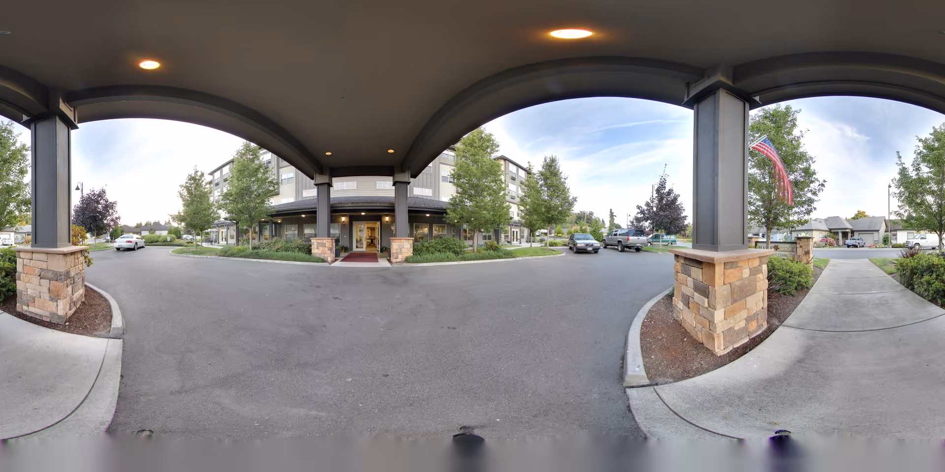 View from under a covered entrance showing the driveway and entrance to a multi-story senior living facility building with stone pillars and landscaped greenery. Several cars are parked nearby, and an American flag is visible on the right side.