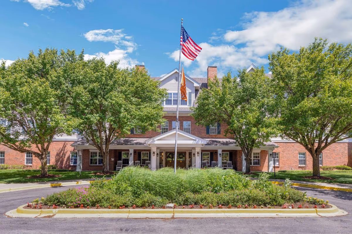Front exterior view of Brighton Gardens of Wheaton, a multi-story brick building with white trim, surrounded by green trees and landscaping. An American flag and another flag are flying on a flagpole in front of the building entrance under a covered porch.