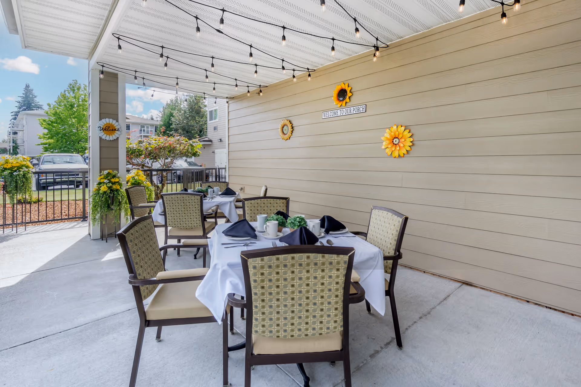 Outdoor covered patio area with a dining table set for four people with white tablecloths, black napkins, and tableware. The patio has string lights hanging from the ceiling and decorative sunflower wall hangings on the beige siding wall. There are plants and flowers around the patio, and a view of neighboring houses and a parked car in the background.