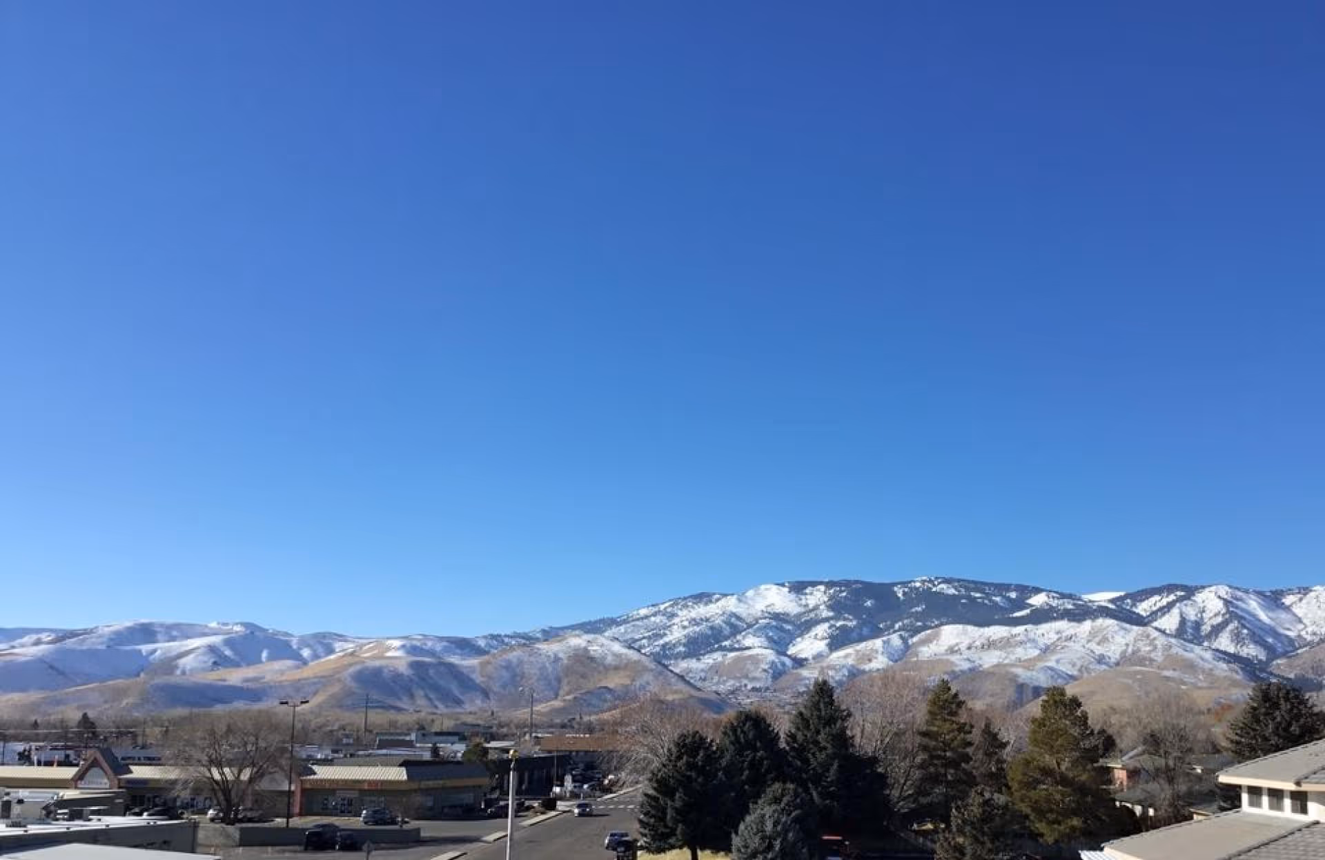 View of snow-capped mountains under a clear blue sky with a small town, trees, and rooftops in the foreground.