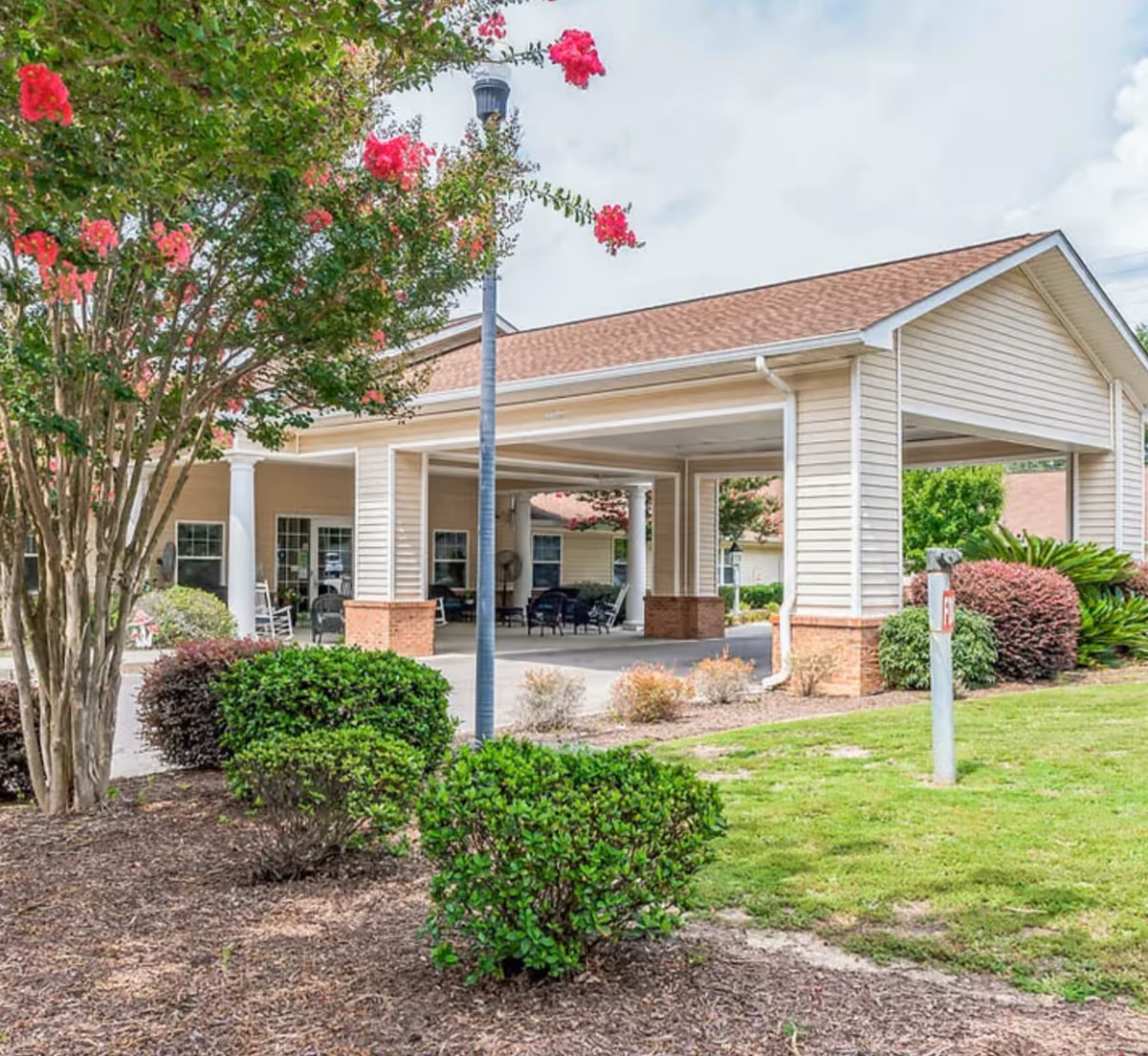 Covered entrance and driveway of a single-story senior living building with columns, outdoor seating, and landscaped shrubs.