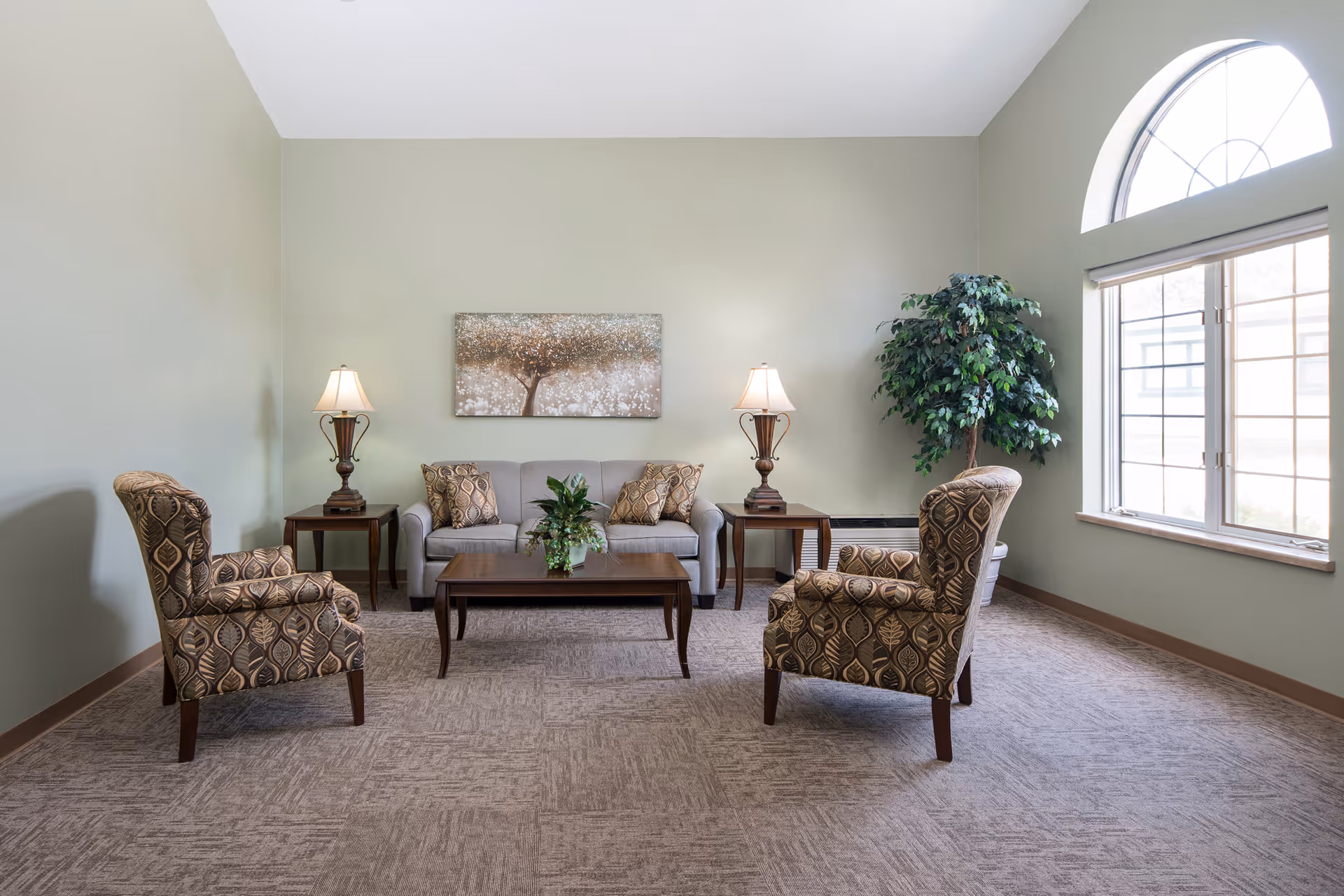 A bright living room with a high ceiling and a large arched window. The room features a gray sofa with patterned cushions, two matching patterned armchairs, a wooden coffee table with a plant centerpiece, two side tables each with a lamp, and a large potted plant in the corner. A decorative painting of a tree hangs on the wall behind the sofa.