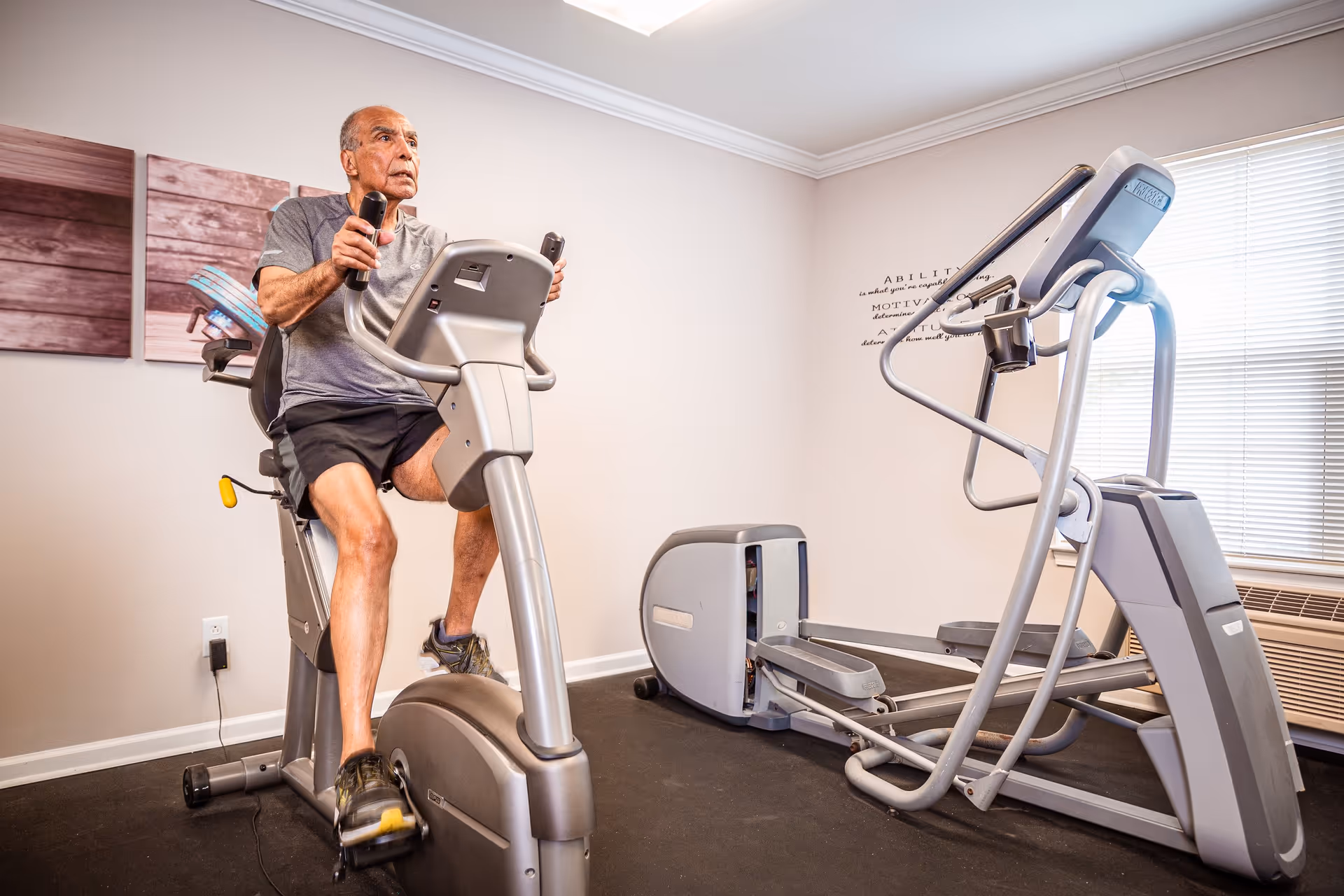 An elderly man exercising on a stationary recumbent bike in a fitness room with another elliptical machine nearby, light-colored walls, and a window with blinds.