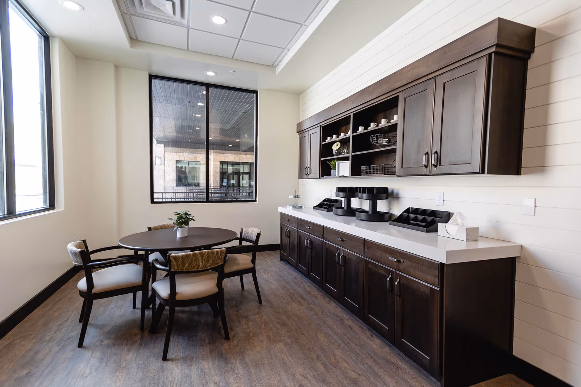 A small dining area with a round table and four cushioned chairs. The room features dark wooden cabinets with white countertops along one wall, holding coffee dispensers, cups, and tissue boxes. Large windows allow natural light to enter the space.