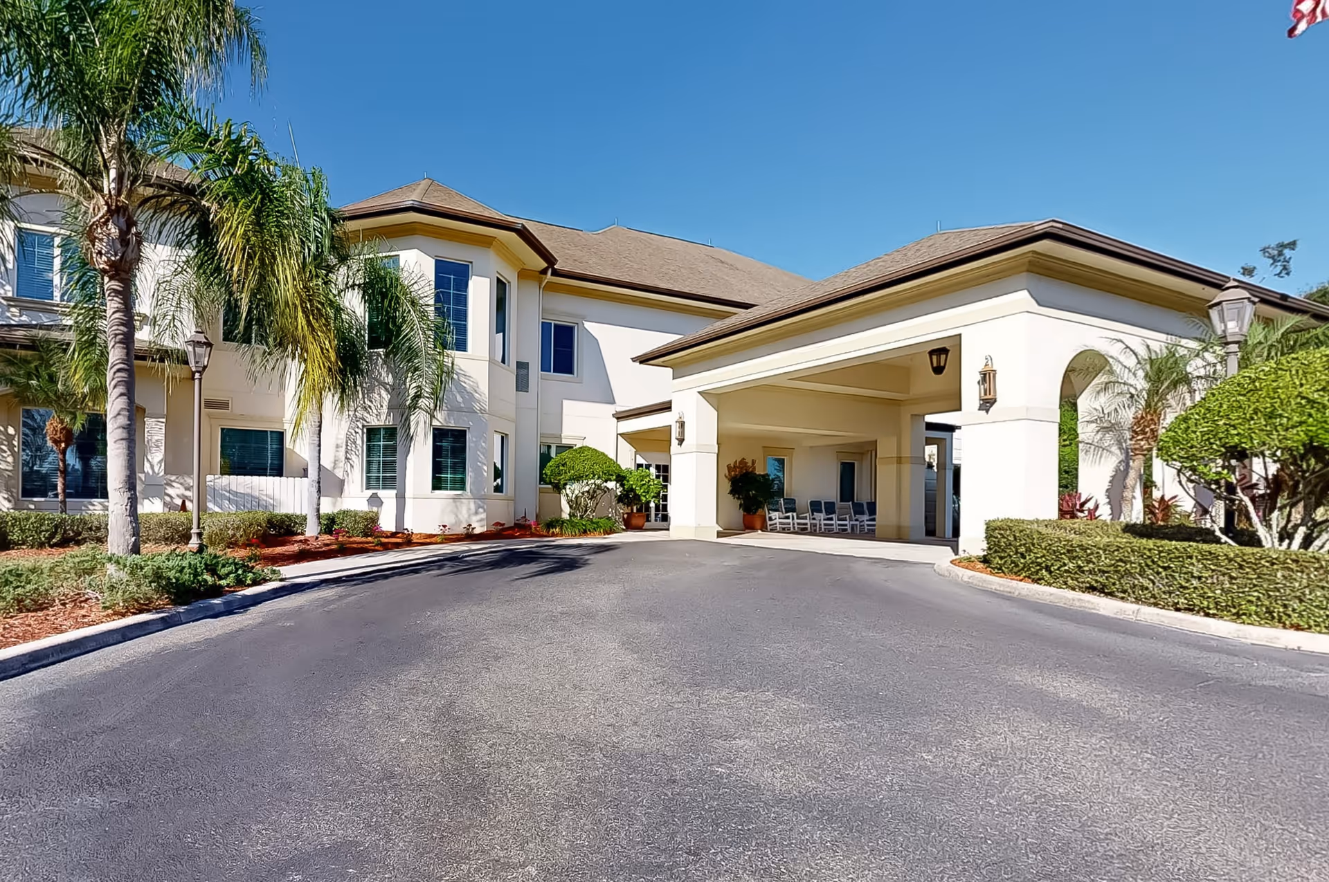Exterior view of The Windsor Of Ocala senior living facility showing a two-story building with a covered entrance, palm trees, and well-maintained landscaping under a clear blue sky.
