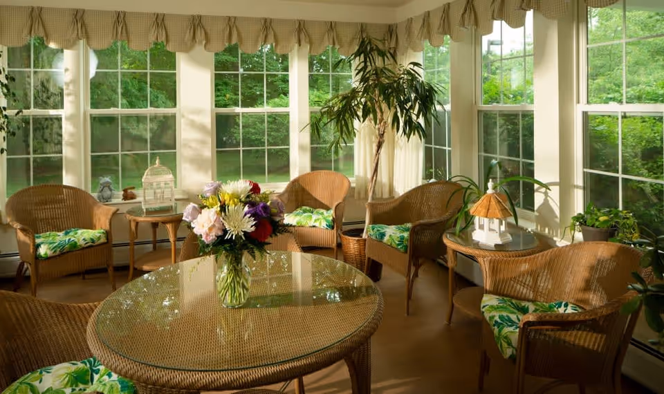 A sunlit room with large windows showing green foliage outside. The room contains wicker furniture including a round glass-top table with a vase of colorful flowers, several wicker chairs with green and white patterned cushions, and side tables with decorative items. The windows have beige valances and the floor is wooden.
