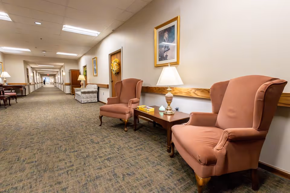 A long, well-lit hallway in an assisted living facility with carpeted floors and beige walls. Along the right side, there are two upholstered armchairs with a wooden side table between them holding a lamp and a box. The walls are decorated with framed pictures and a wreath on a door. Additional seating and lamps are visible further down the hallway.