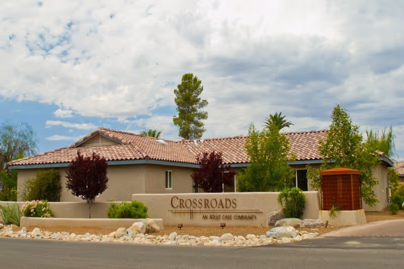 Exterior view of Crossroads Assisted Living facility showing a single-story building with a tiled roof, surrounded by landscaping including trees, bushes, and rocks. A sign in front reads 'Crossroads An Adult Care Community'. The sky is partly cloudy.