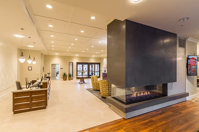 Interior view of a senior living facility lobby with a modern double-sided fireplace, a wooden reception desk on the left, seating area with patterned chairs, and entrance doors in the background.
