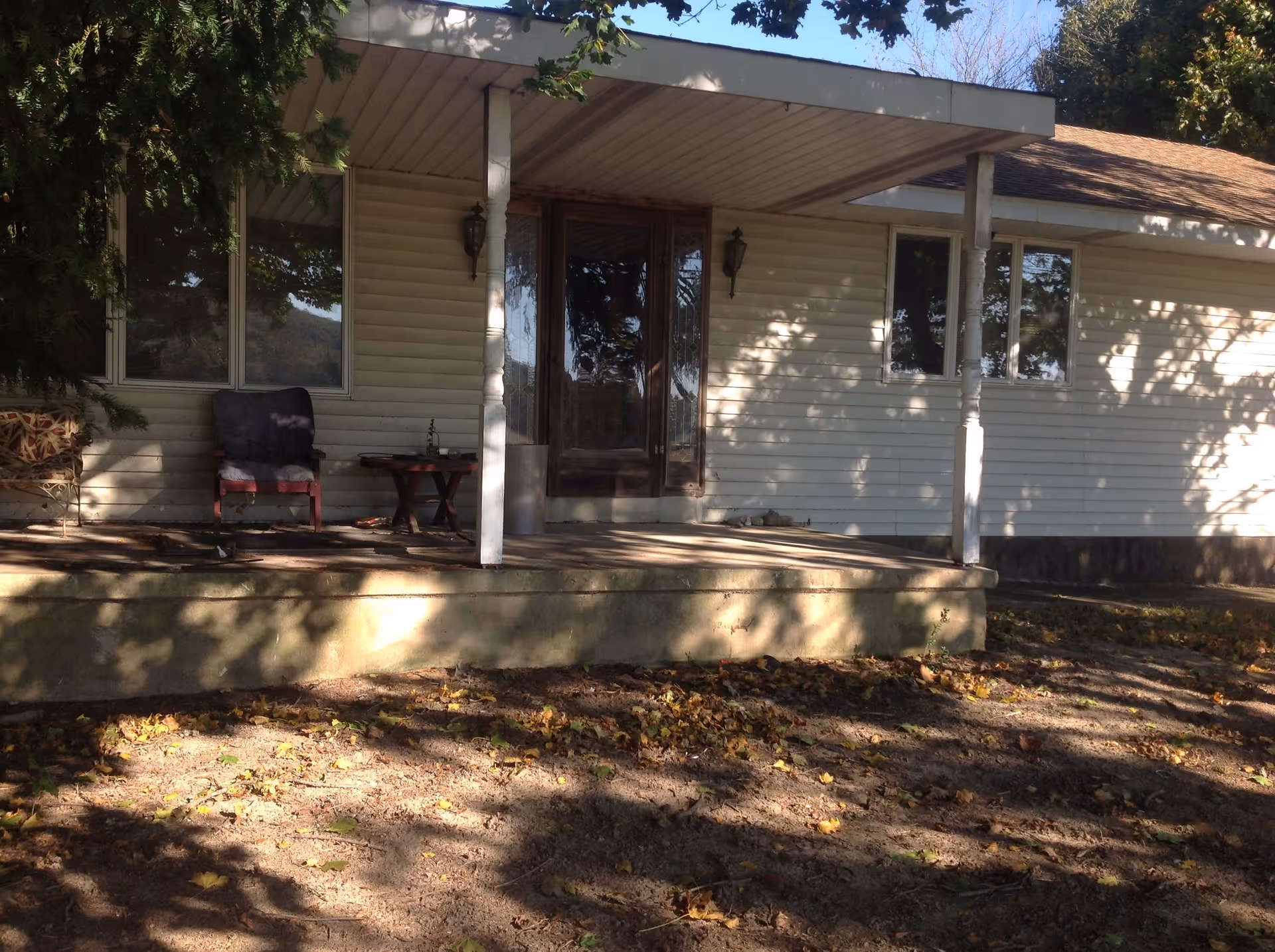 Front porch of a single-story house with beige siding, two white pillars supporting a roof over the porch, a wooden door with glass panels, two wall-mounted lantern lights, a cushioned chair, a small table, and a bench. The ground in front of the porch is covered with dry leaves and dirt, and trees cast shadows on the house and ground.
