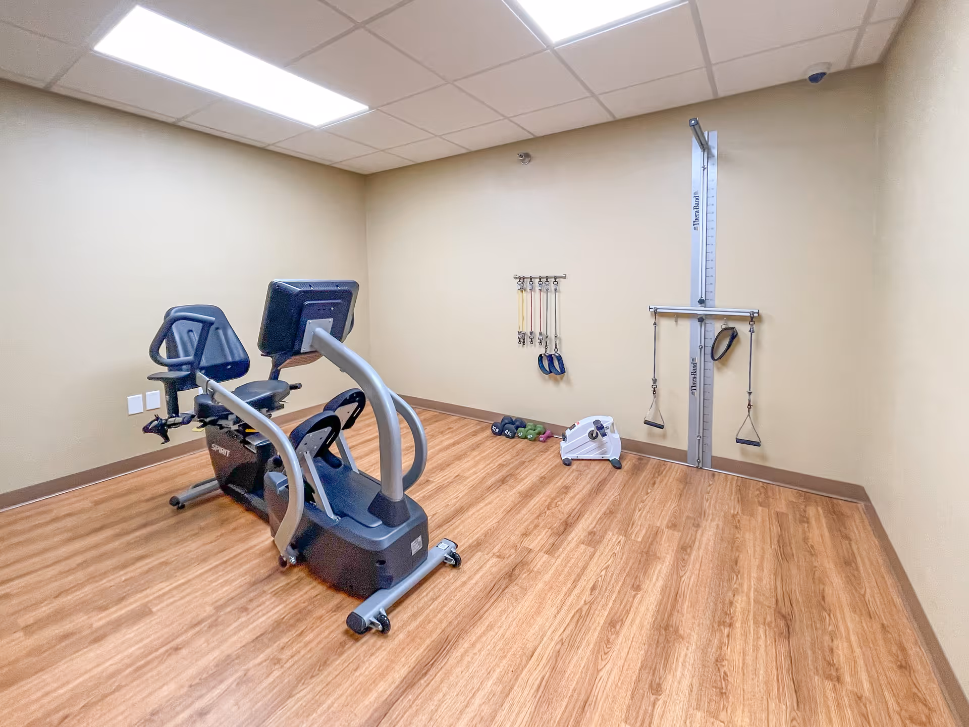 A small exercise room with light wood flooring and beige walls. The room contains a recumbent exercise bike, a wall-mounted pulley system for resistance training, several resistance bands hanging on the wall, a set of small dumbbells, and a pedal exerciser on the floor. The ceiling has recessed lighting panels.