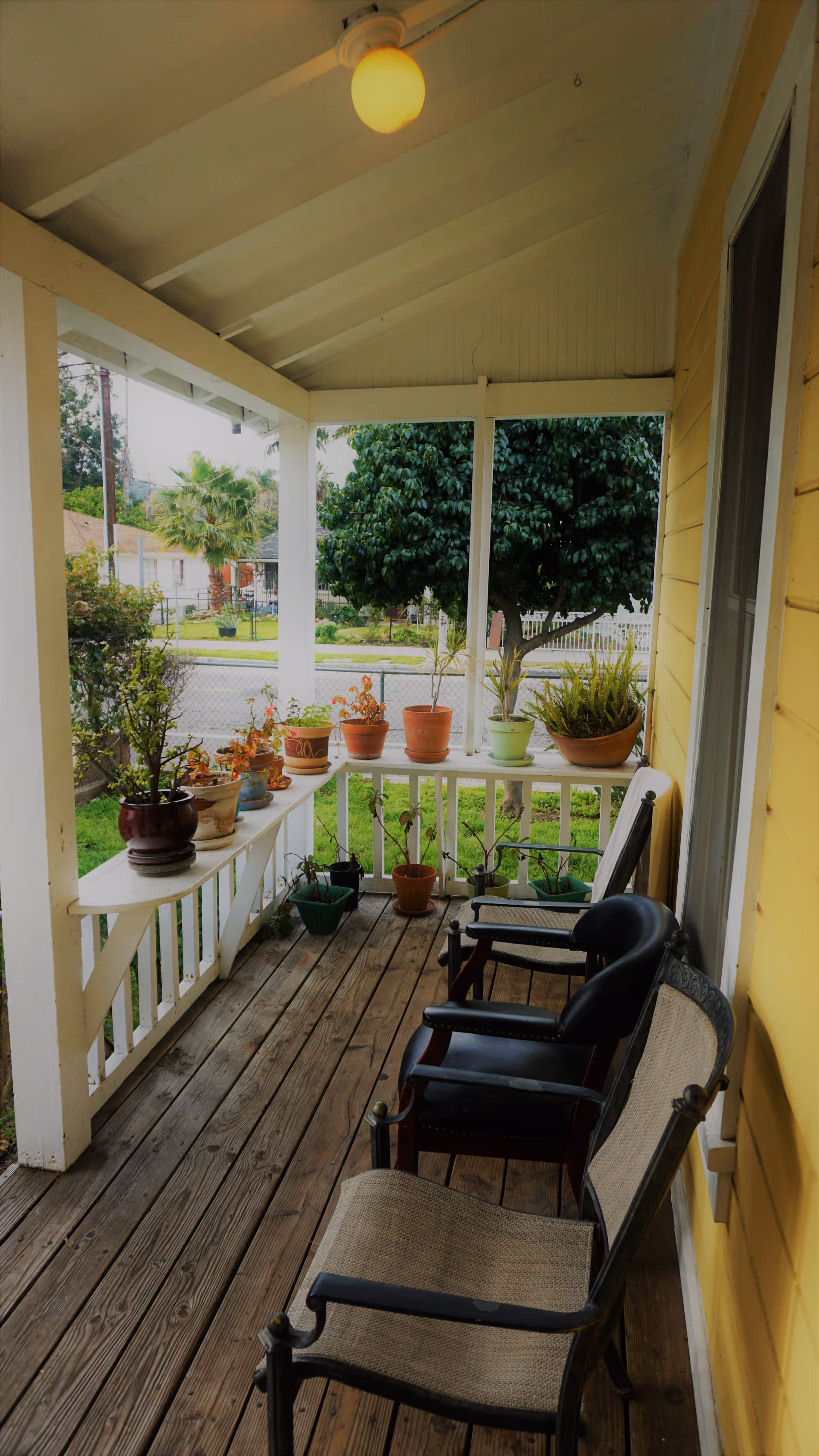 A covered wooden porch with three chairs and several potted plants arranged on the railing and floor. The porch has a white ceiling with a round light fixture and yellow exterior walls. In the background, there is a view of a street, trees, and neighboring houses.