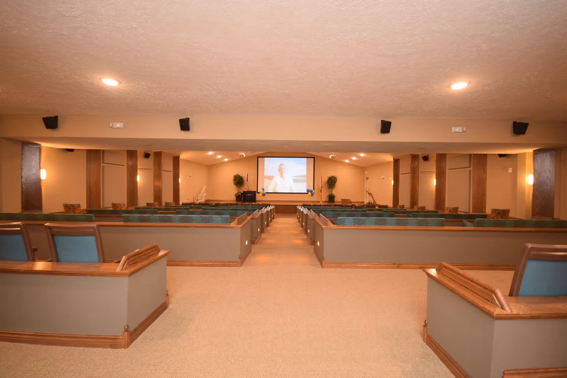 Interior view of a large room with rows of green cushioned pew-style seating facing a screen at the front displaying a person. The room has beige walls, carpeted floor, wooden trim, and soft lighting with wall sconces and ceiling lights.