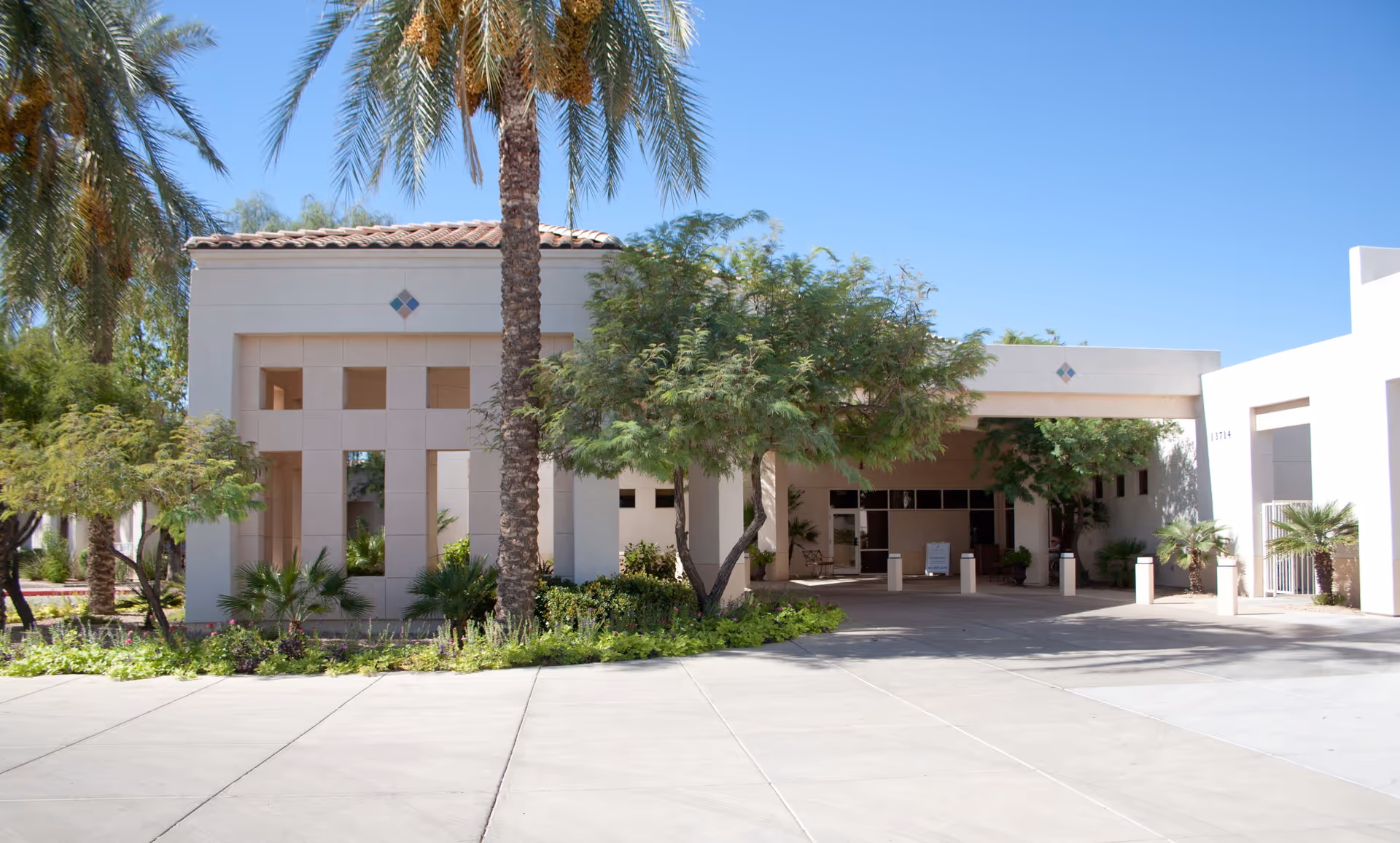 Exterior view of Casa del sol facility entrance with a covered driveway, surrounded by palm trees and other greenery under a clear blue sky.