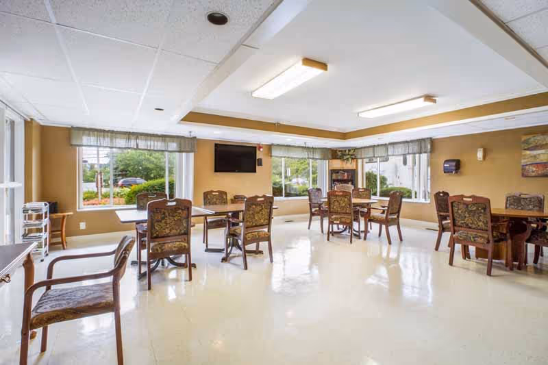 Bright communal dining room with several tables and upholstered chairs arranged near large windows.