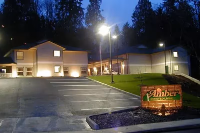 Exterior view of The Amber assisted living facility at dusk, showing a two-story building with illuminated windows and outdoor lights, a parking lot with marked spaces, and a lit sign reading 'Amber Assisted Living' on a brick base in the foreground.