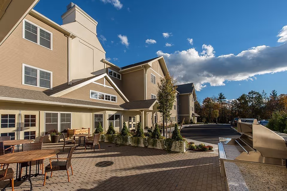 Sunlit exterior patio with tables, potted plants, and a multi-story senior living building under a blue sky.