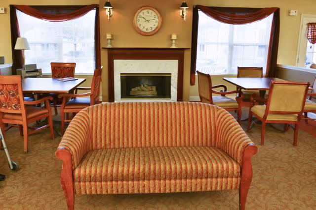 A cozy living room area with a red and gold striped sofa in the foreground, two windows with red curtains, a fireplace with a clock above it, and two tables with chairs arranged around them.