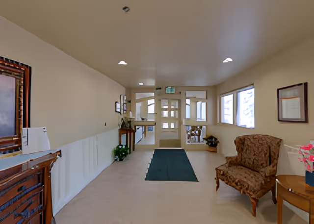 A hallway in an assisted living facility with a green carpet runner leading to a glass door exit. The hallway has beige walls and ceiling with recessed lighting. On the right side, there is a patterned armchair next to a small round table with a pink flower arrangement. On the left side, there is a wooden console table with framed artwork above it and a small plant below.