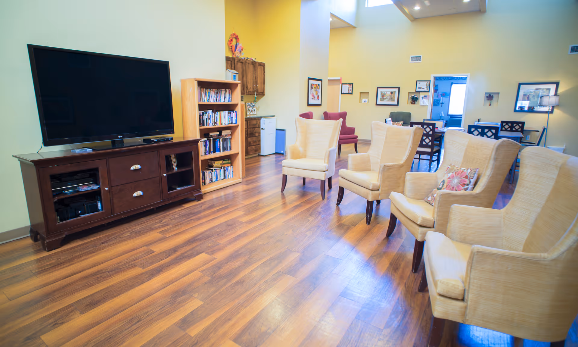 Bright communal living room with a large TV, bookshelf, and a row of upholstered armchairs on wood floors.