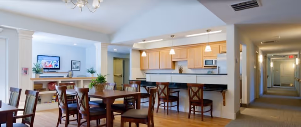 Open communal dining area and kitchenette with wooden tables and chairs, bar seating, TV and adjacent hallway.