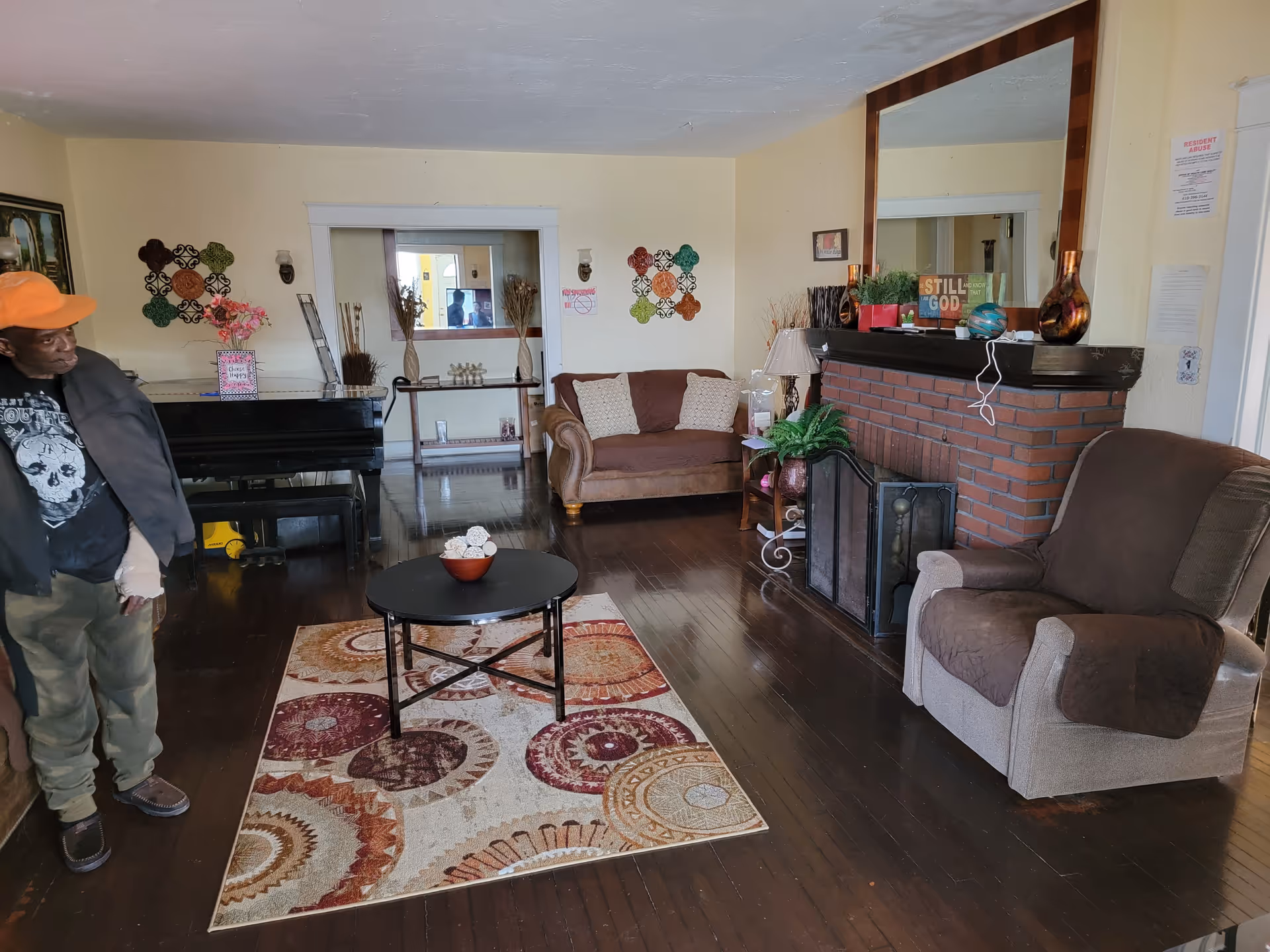 A cozy living room with dark wooden floors, a patterned area rug, a round black coffee table with decorative items, a brown loveseat, and a beige armchair with a brown cover. There is a brick fireplace with a large mirror above it and various decorative items on the mantel. A black piano is against the wall on the left side, and a man wearing an orange cap and dark jacket is standing near it. The walls are light-colored with some wall decorations and framed pictures.