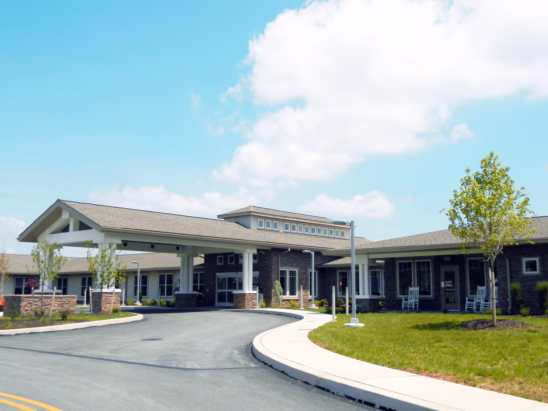 Exterior view of a single-story senior living facility building with a covered entrance driveway, landscaped lawn, young trees, and a clear blue sky with some clouds.