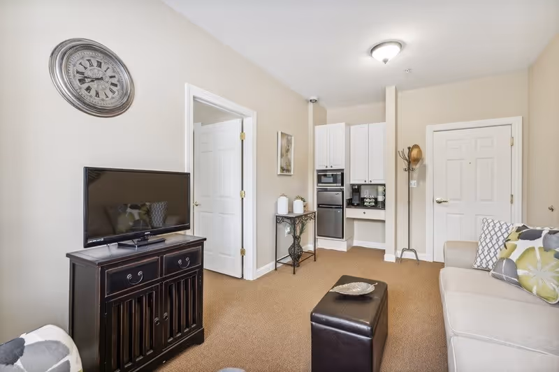 A cozy living room with beige walls and carpet. There is a beige sofa with patterned cushions on the right, a dark wooden TV stand with a flat-screen TV on the left, and a small black ottoman in the center. A round wall clock hangs above the TV. In the background, there is a small kitchenette area with white cabinets, a built-in microwave, and a coffee maker. A coat rack with a hat is near a white door, and a small decorative table with plants and artwork is against the wall.