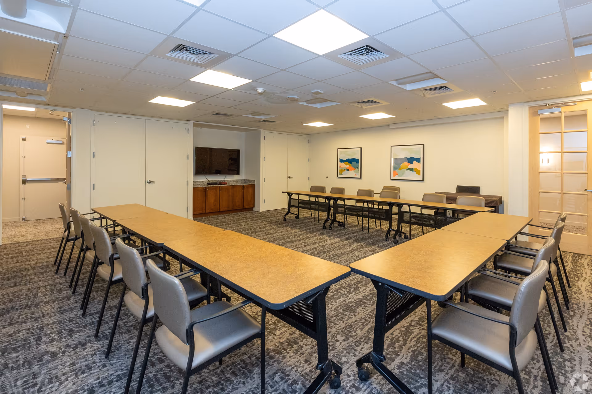 A conference or meeting room with tables arranged in a U-shape surrounded by chairs. The room has a carpeted floor, a ceiling with recessed lighting, a wall-mounted TV, two abstract paintings on the wall, and a wooden cabinet beneath the TV. There is a door with glass panels on the right side and a closed door at the back.