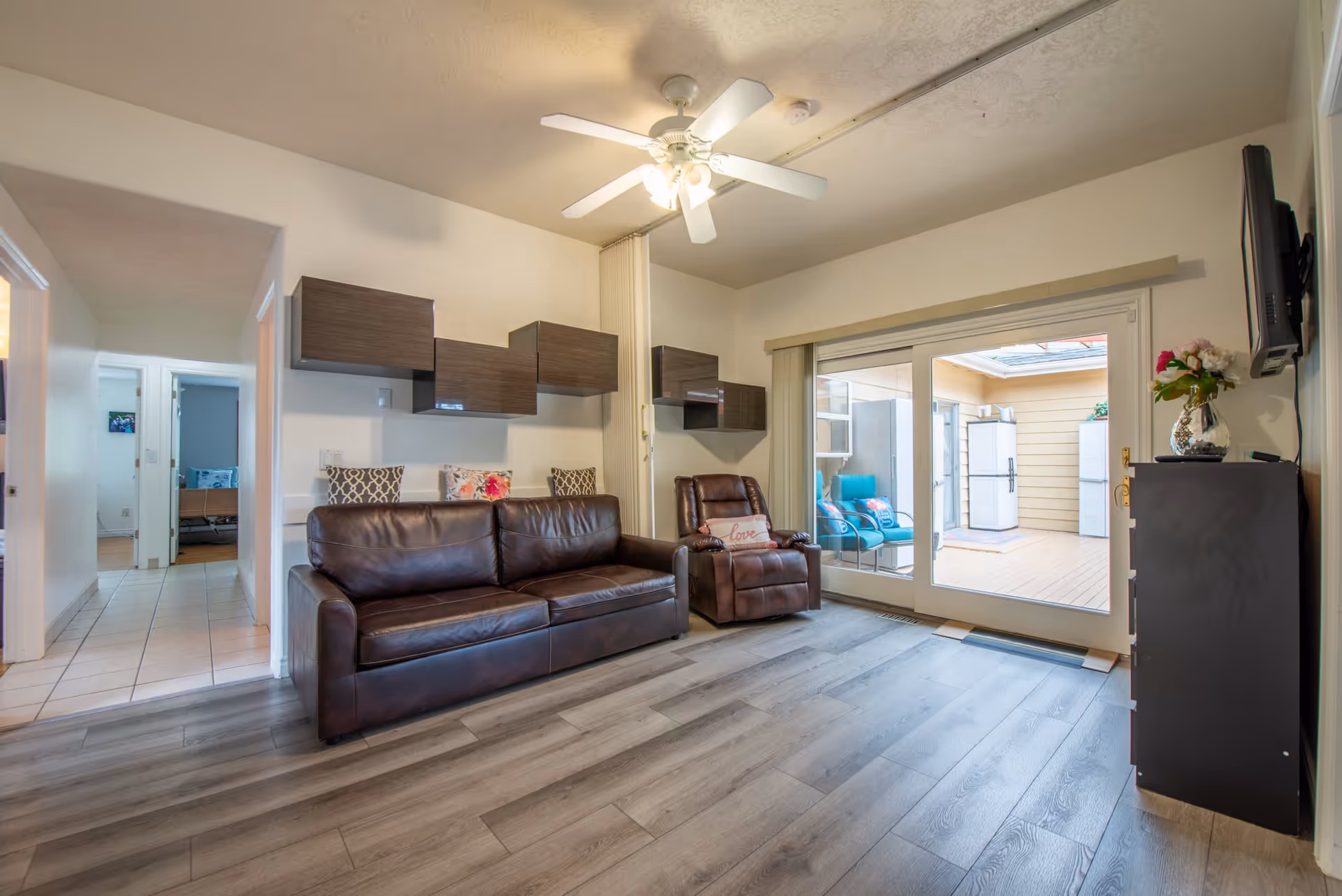 Living room with a brown leather sofa and recliner, ceiling fan, wall-mounted TV, and sliding glass doors opening to a sunroom.