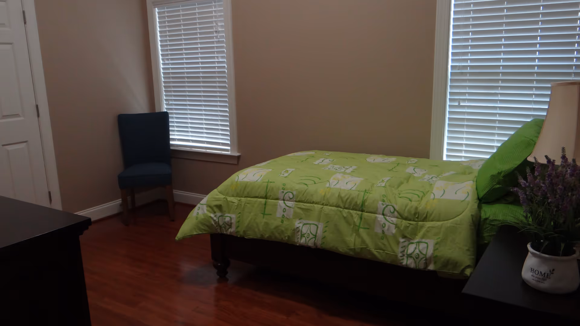 A bedroom with a single bed covered in a green patterned comforter and matching pillow. There is a dark wooden nightstand next to the bed with a lamp and a small potted plant. A blue upholstered chair is placed near a window with white blinds. The room has wooden flooring and beige walls.