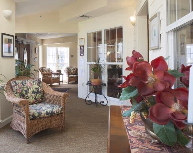 Interior view of a senior living facility lounge area with wicker chairs featuring floral cushions, a small table with a potted plant, large windows, and a decorative arrangement of red flowers in the foreground.