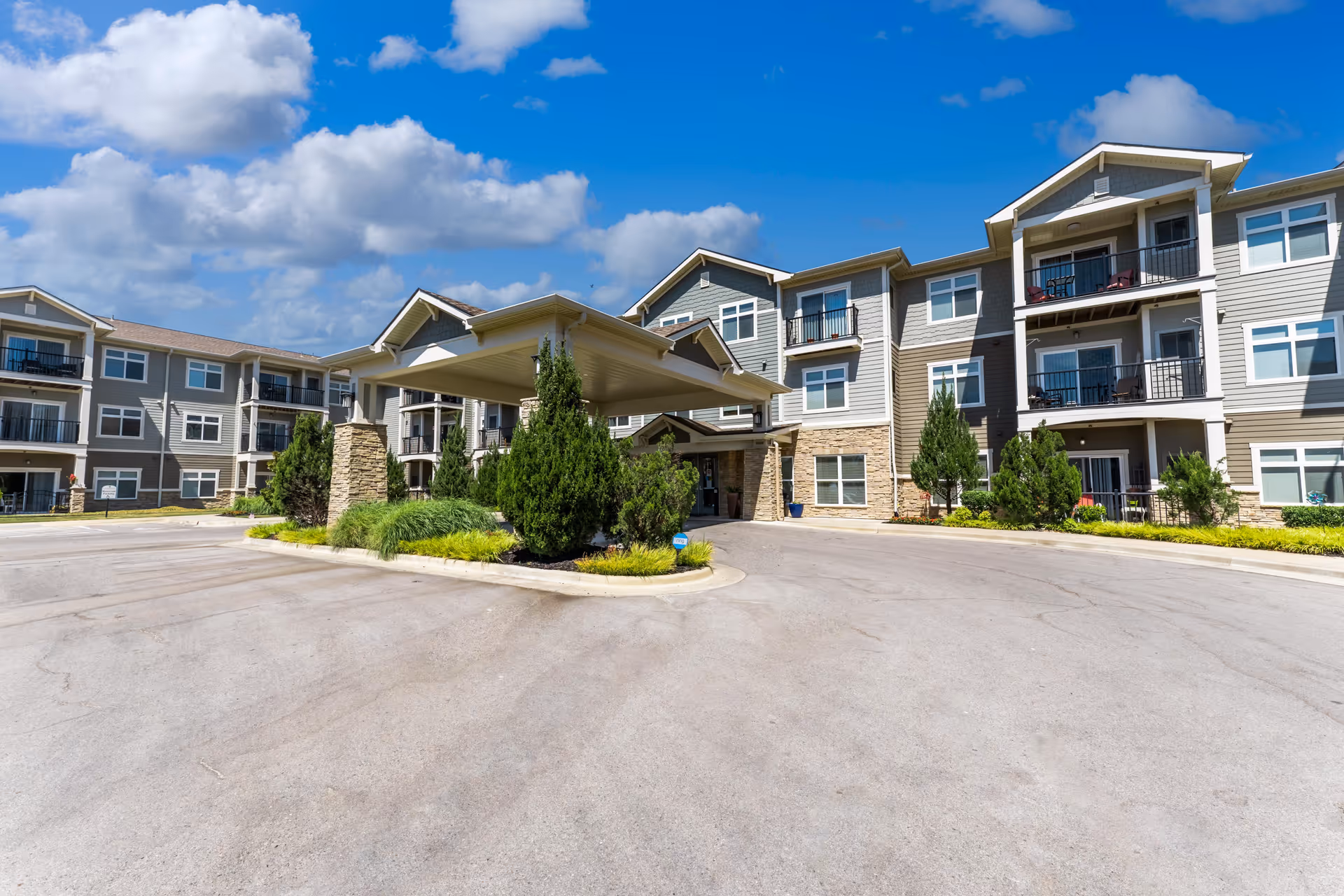 Exterior view of a senior living facility named The Cedars at Woodland Hills, featuring a three-story building with balconies, a covered entrance with stone pillars, landscaped greenery, and a clear blue sky with some clouds.