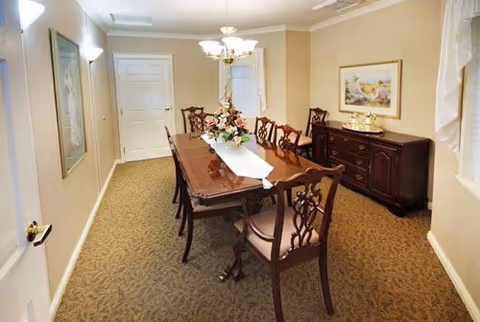 A formal dining room with a long wooden dining table surrounded by eight matching chairs. The table is decorated with a white runner and a floral centerpiece. The room has beige walls, carpeted floor, a chandelier overhead, a sideboard with decorative items, and framed artwork on the walls.
