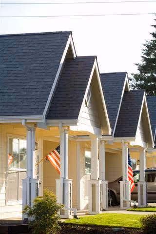 Row of small residential-style units with peaked roofs and front porches, each displaying an American flag, with a well-maintained lawn and trees in the background.