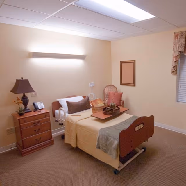 A small, neatly arranged bedroom in a healthcare facility featuring a single hospital bed with beige bedding and pillows. Next to the bed is a wooden nightstand with a lamp, a phone, and a small plant. A cushioned chair with a pillow is placed against the wall, and a window with a valance is visible on the right side. The room has beige walls and carpeted flooring with a ceiling light panel.
